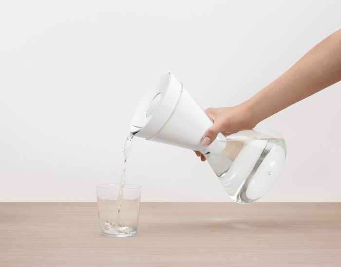 Person pouring water from a soma carafe into a glass on a light wooden surface.