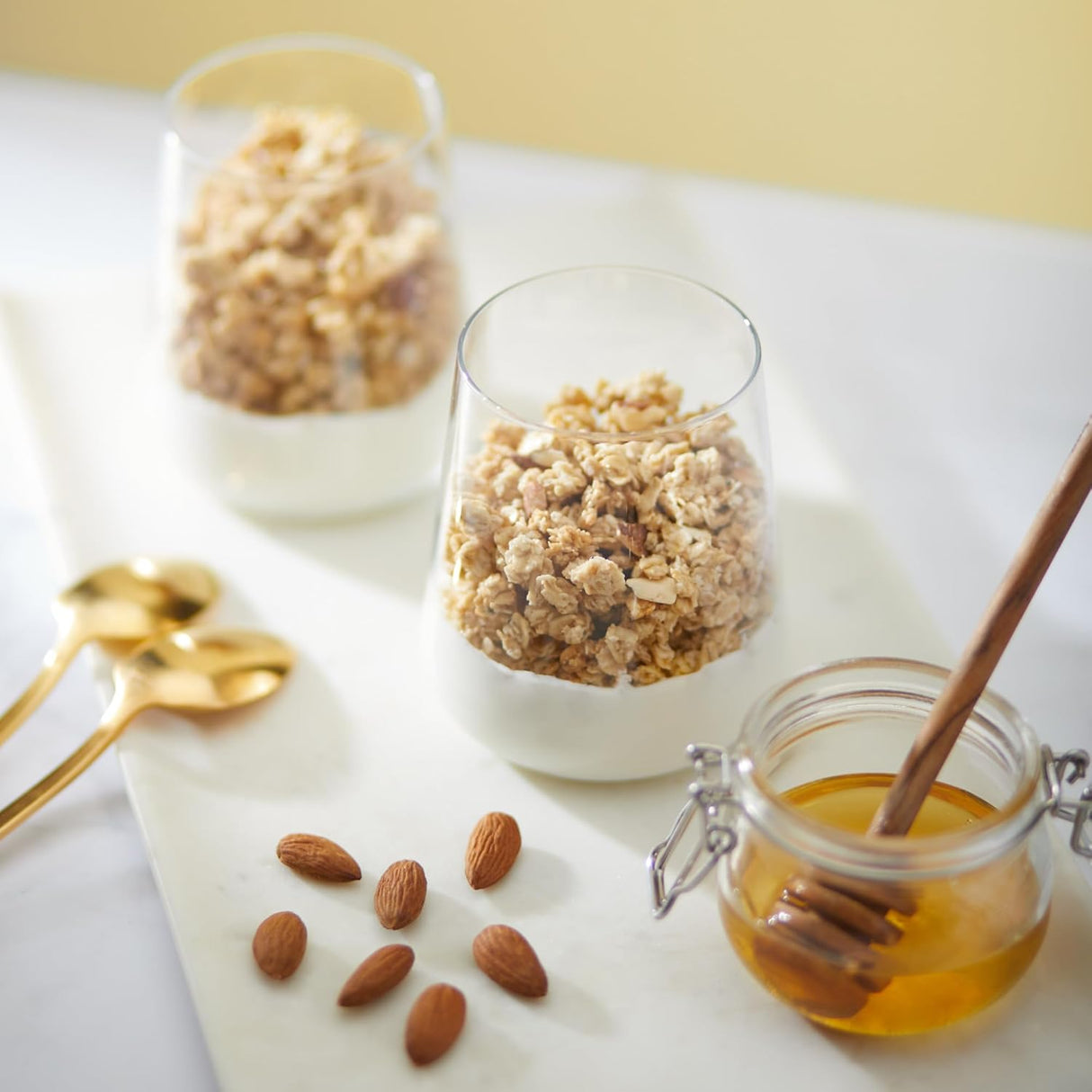 Two glasses of granola with a jar of honey and almonds on a white surface.