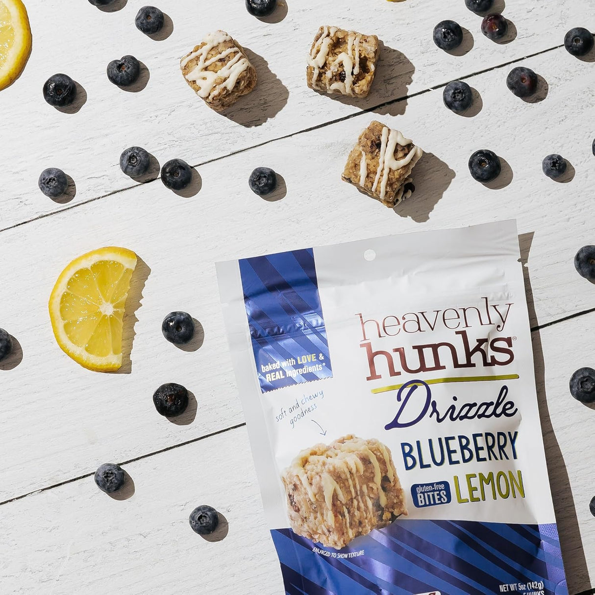 Packaging of Heavenly Hunks Drizzle Blueberry Lemon bites with lemon slices and blueberries on a white surface.