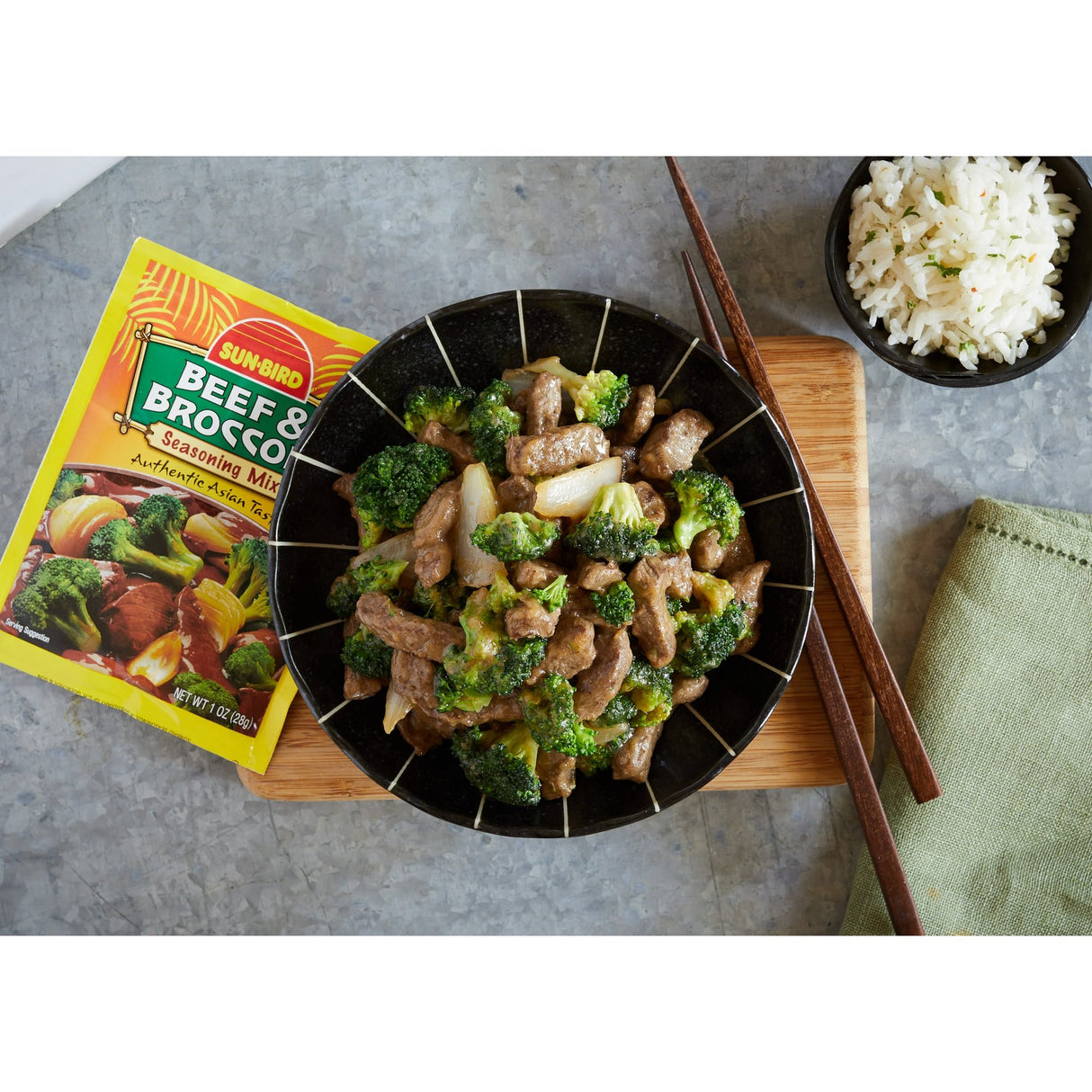 Beef and broccoli stir-fry in a black bowl with Sunbird seasoning mix on a gray surface.