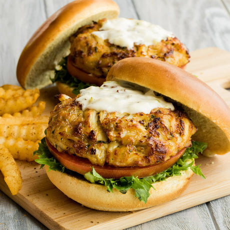 Two crab cake burgers with lettuce and sauce on buns, served with fries on a wooden board.
