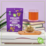 Purple package of Organic Sprouted Snacking Seeds with a glass of tea and bowl on a white surface.