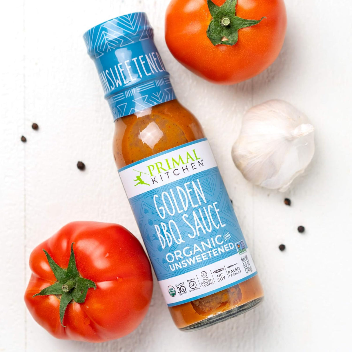 Bottle of Primal Kitchen Golden BBQ Sauce with tomatoes and garlic on a white background