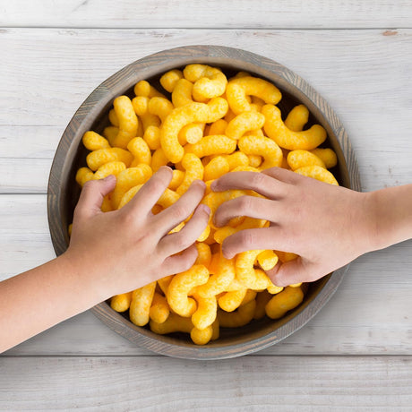 Two hands reaching into a bowl filled with yellow snack sticks on a wooden surface.