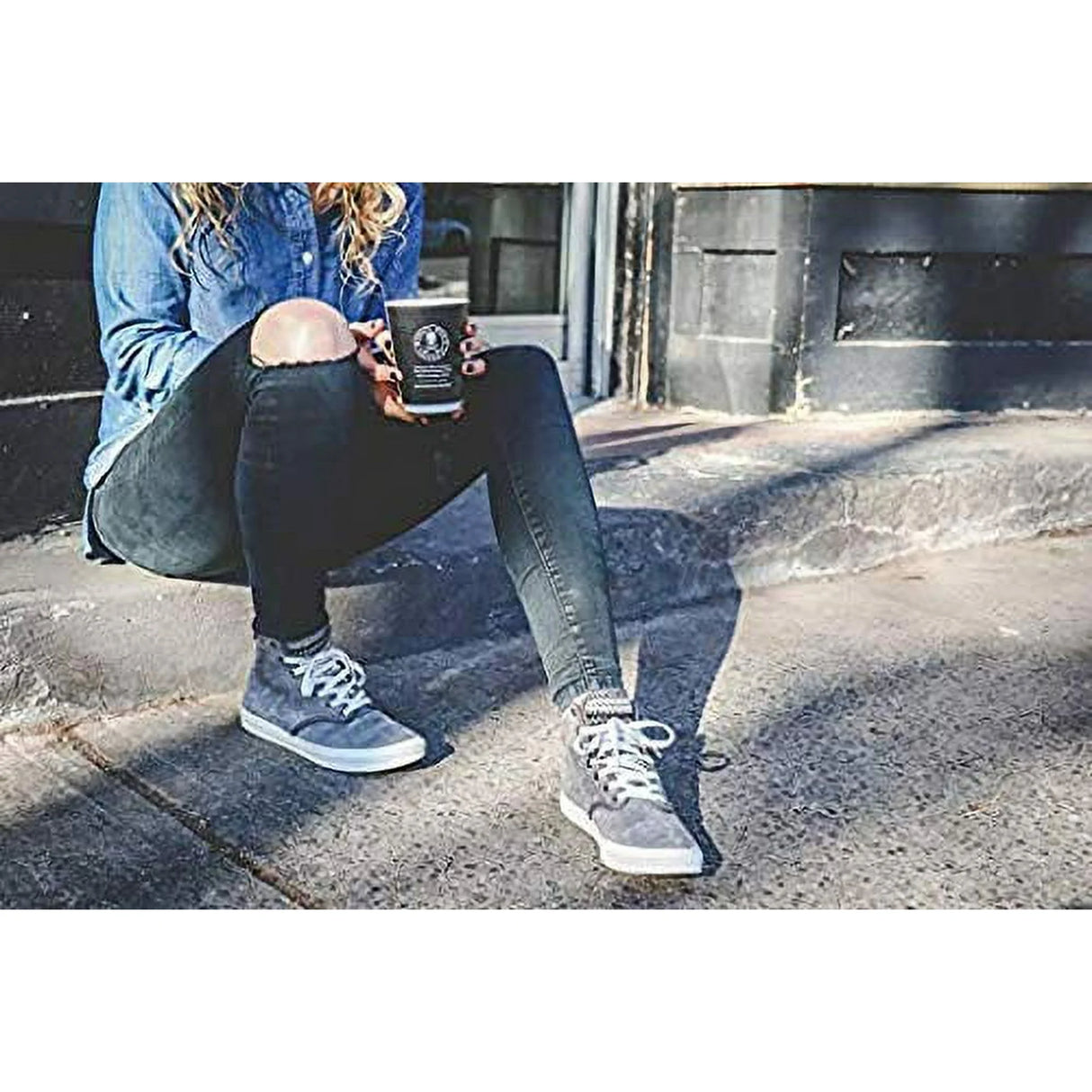 Person sitting on steps holding a coffee cup, wearing jeans and sneakers.