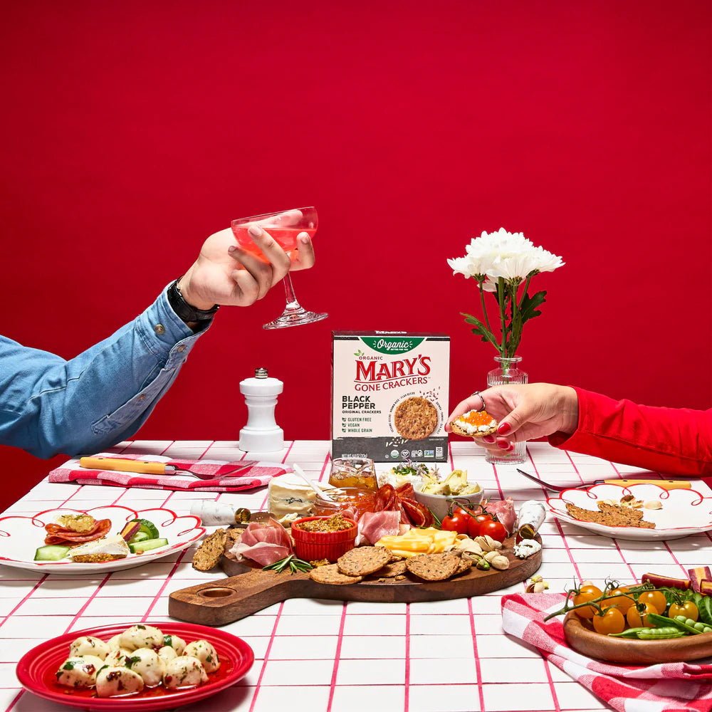 Two people at a table with a spread of food, drinks, and a box of Mary's Gone Crackers against a red background.