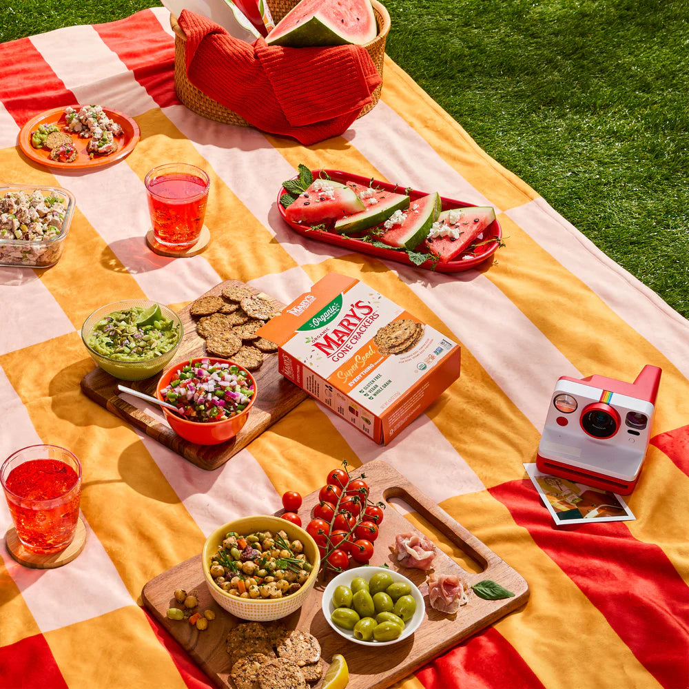 Outdoor picnic setup with food, drinks, and a camera on a colorful blanket.