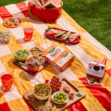 Outdoor picnic setup with food, drinks, and a camera on a colorful blanket.