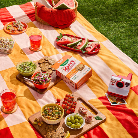 Outdoor picnic setup with food, drinks, and a camera on a colorful blanket.