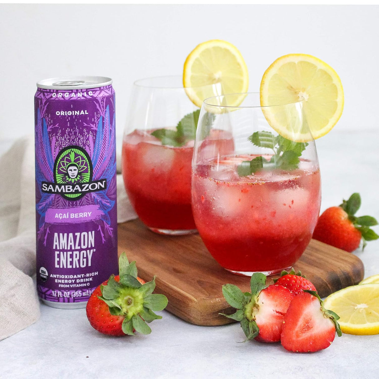 Sambazon Amazon Energy drink can with two glasses of red juice garnished with lemon slices and strawberries on a white background.