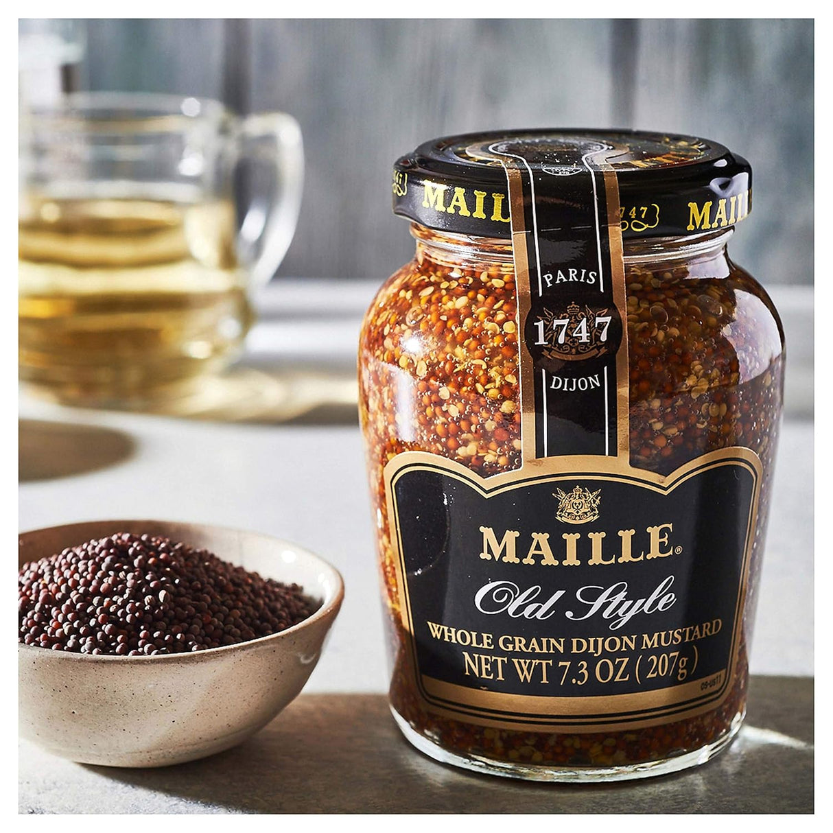 Jar of Maille Old Style Whole Grain Dijon Mustard with a bowl of mustard seeds on a table.