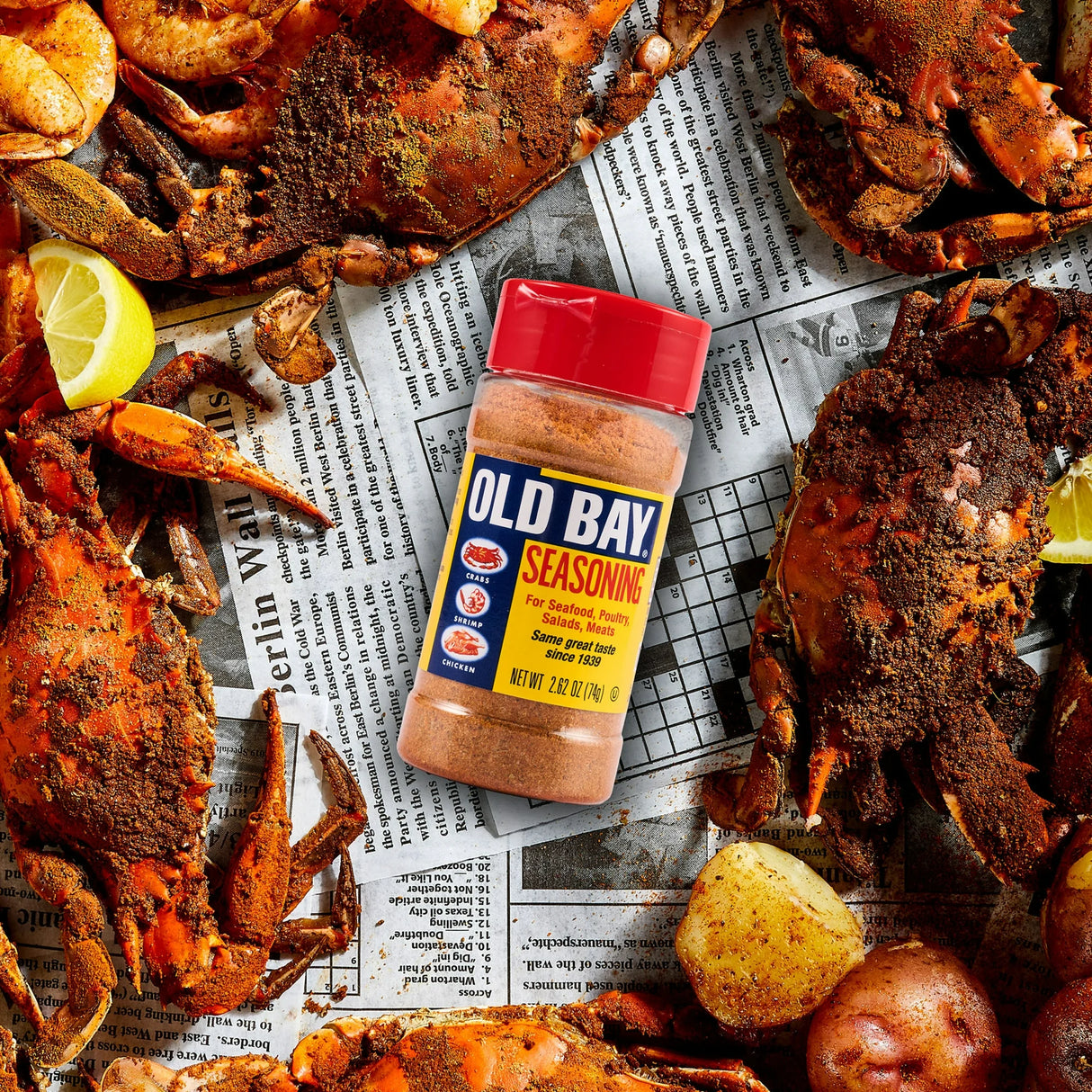 Old Bay seasoning bottle surrounded by crabs and potatoes on a newspaper background