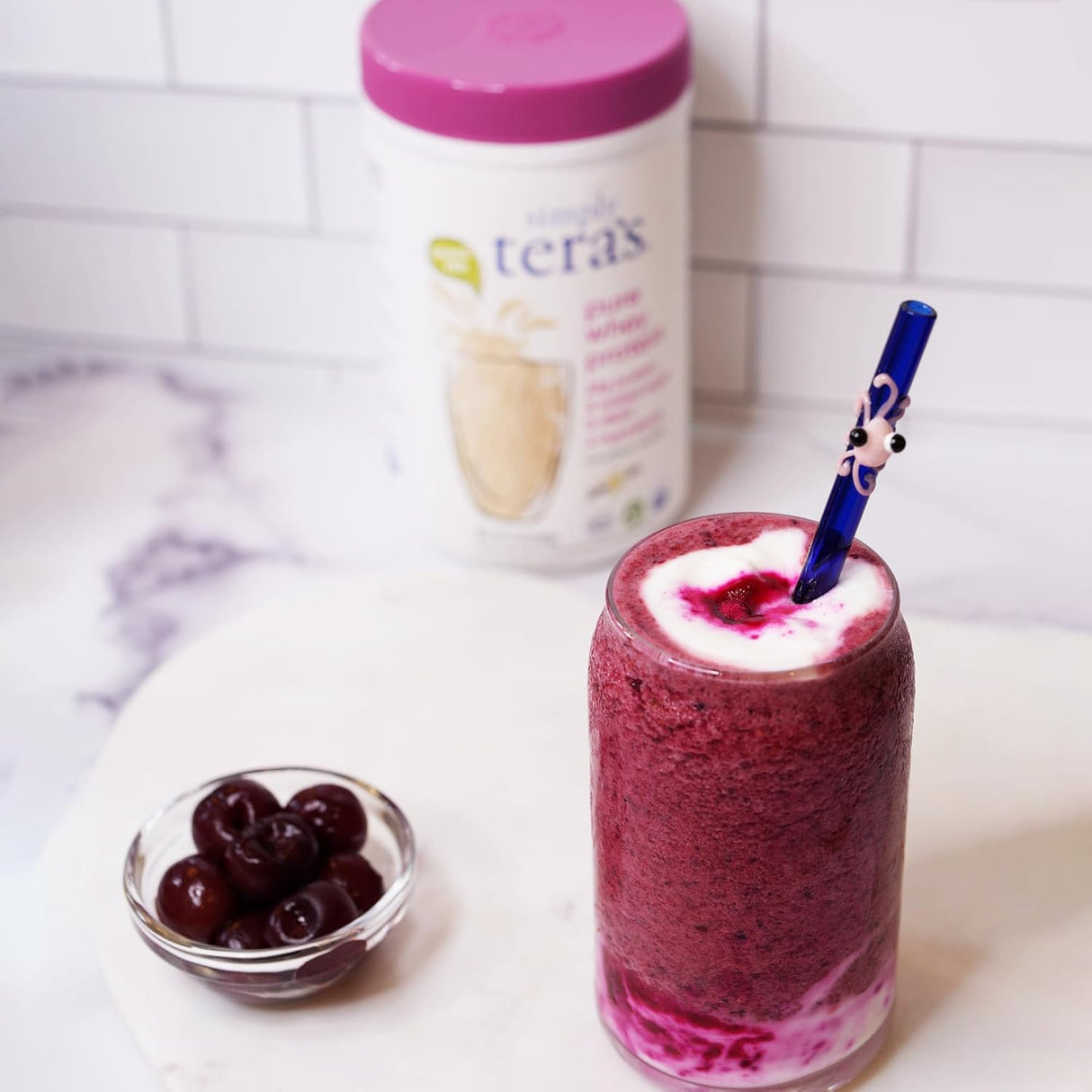Glass of red smoothie with a blue straw, container of yogurt, and bowl of cherries on a white surface.