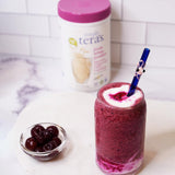 Glass of red smoothie with a blue straw, container of yogurt, and bowl of cherries on a white surface.