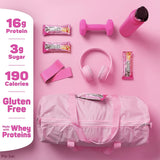 Pink gym bag with fit crunch protein bars, dumbbells, headphones, and a water bottle on a pink background.