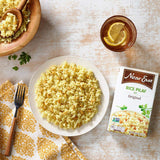 Plate of rice pilaf with a box of Near East Rice Pilaf, a bowl of rice, and a glass of lemonade on a wooden table.