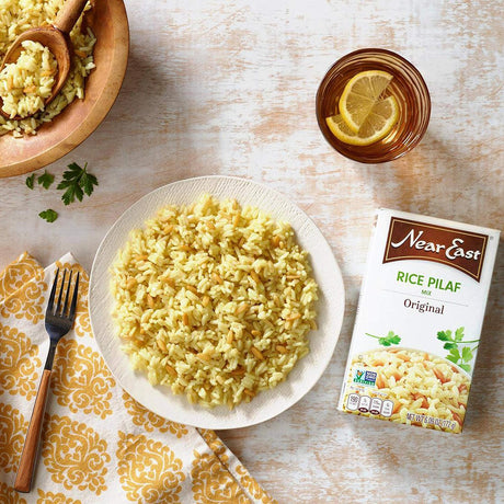 Plate of rice pilaf with a box of Near East Rice Pilaf, a bowl of rice, and a glass of lemonade on a wooden table.