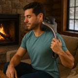 A man uses a handheld massager on her neck while sitting on a cozy couch near a stone fireplace.